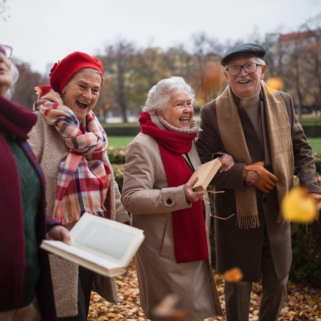 A group of happy senior friends with books on walk outdoors in park in autumn, talking and laughing